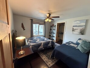Une chambre à coucher comprenant un lit, une table de chevet, une lampe, un ventilateur de plafond, une fenêtre, une porte, une étagère avec des objets décoratifs et un tableau au mur.