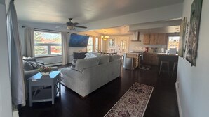 Amplia sala de estar con sofá, televisor y mesa de comedor. Se ve una cocina con armarios de madera y estufa a gas.