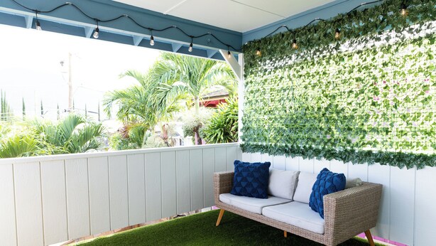 Une terrasse couverte avec une clôture blanche, une pelouse artificielle verte, un canapé en osier avec des coussins bleus et un mur décoré de feuillage vert.