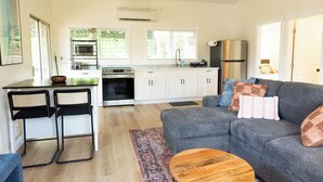 Une cuisine moderne avec des armoires blanches, un four encastré et un réfrigérateur en acier inoxydable. Le salon est aménagé avec un canapé gris doté de coussins et une table basse en bois.