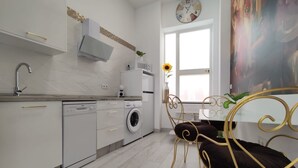 A modern kitchen with white appliances, a built-in oven, and a dining area with a metal chair and table. - The Urban Elegnace Suite (Sevilla)