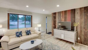 Un salon avec un canapé beige, une table basse, un téléviseur posé sur un meuble en bois et une fenêtre avec des stores.