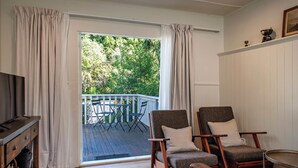 Ein Zimmer mit Holzboden, zwei Sesseln, einem Fernseher und Blick auf einen Balkon mit Gartenmöbeln.