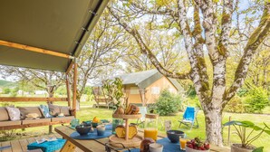 An outdoor dining area with a wooden table set for breakfast, surrounded by greenery and a tented structure.