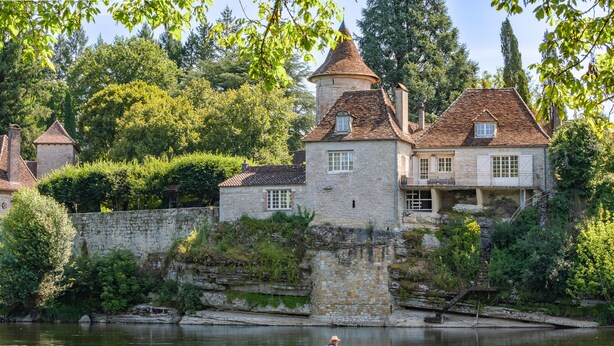 A historic stone building with a tower, situated by a calm river with a person kayaking.
