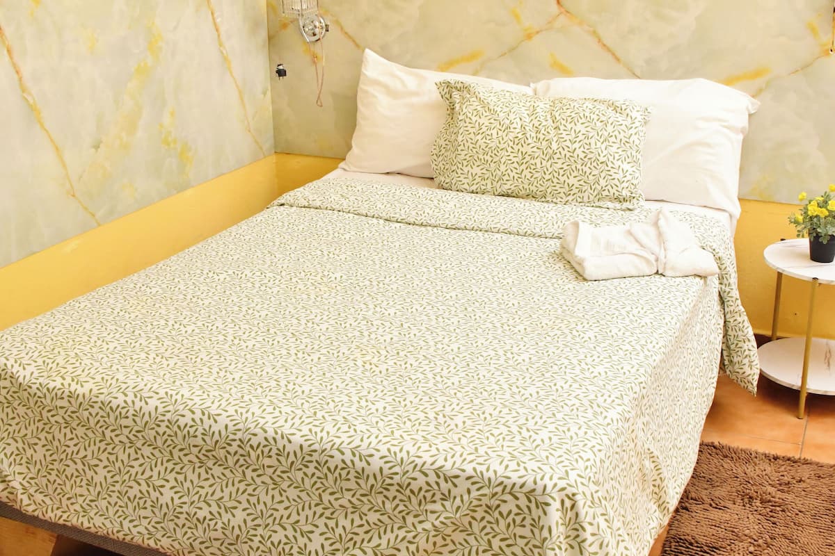 A bed with a leaf-patterned bedspread, a small bedside table with a plant, and a marble-patterned wall.