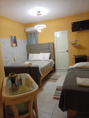 Une chambre d’hôtel de taille modeste, comprenant un lit, une petite table avec un vase, un téléviseur fixé au mur et une porte.