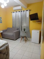 Una habitación con un sofá gris, una mesita con una planta, un refrigerador blanco y un televisor montado en la pared.