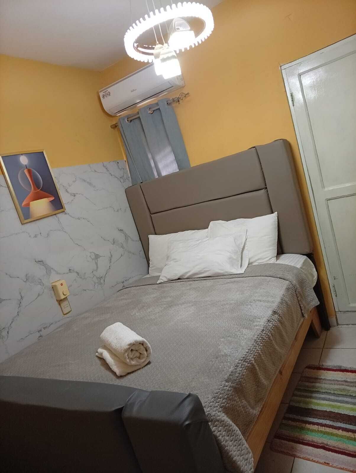 Une chambre d’hôtel avec un lit, une table de chevet, une serviette, un climatiseur fixé au mur, une porte et un tableau encadré accroché au mur.