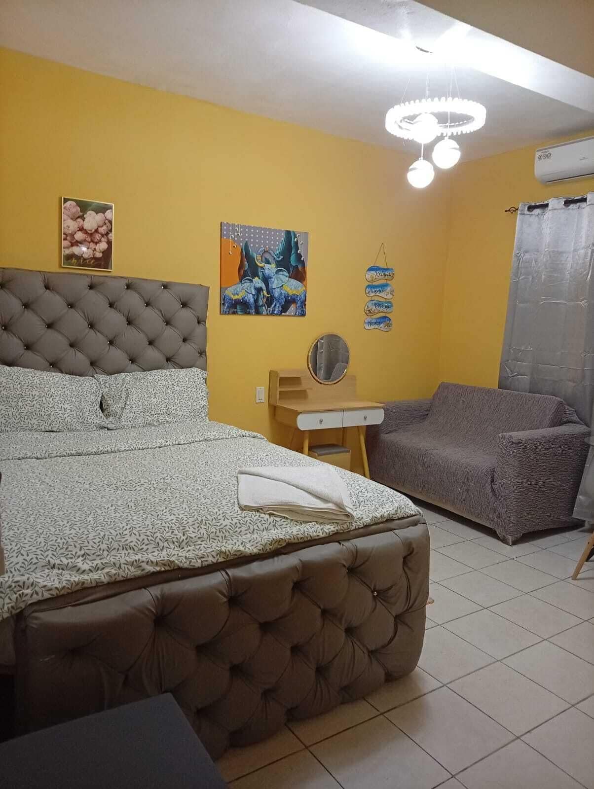 A bedroom with a large bed, a gray sofa, a small table, and a mirror.