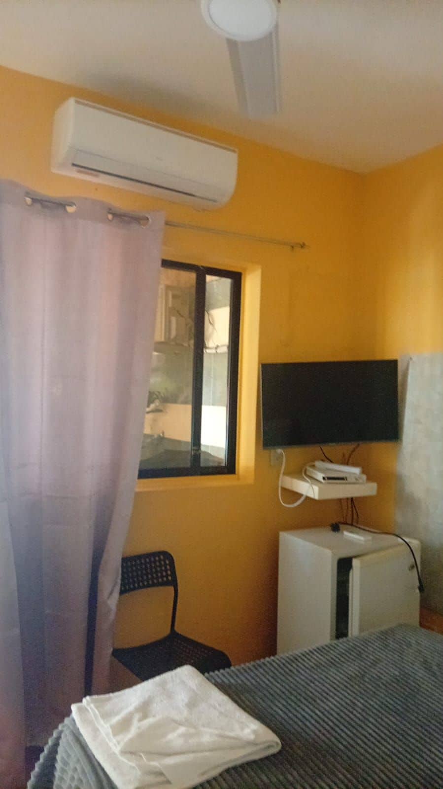 A room with a bed, a desk, a chair, an air conditioning unit, a television, and a window with a curtain.