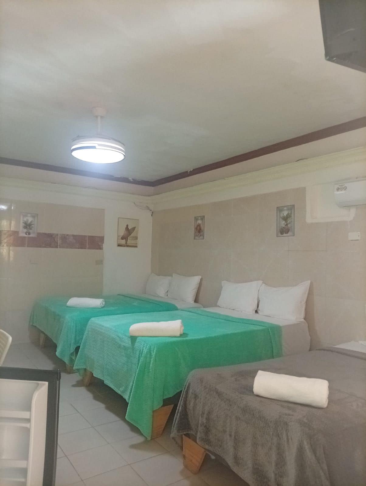 A room with two beds, each with a green bedspread and white pillows, and a grey bedspread with a white pillow. There is a ceiling light fixture and an air conditioning unit on the wall.