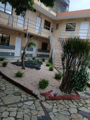A multi-story building with numbered doors, a staircase, and a small garden area with plants and gravel. - Hotel Condo Mango (Boca Chica)