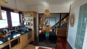 Una cocina moderna con armarios de madera, encimera negra y una isla central con taburetes. Hay una zona de comedor con una silla de mimbre y una escalera que conduce a otro nivel.