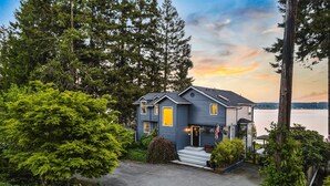 A two-story house with a grey exterior, a front porch, and a view of a lake. - Puget Sound Waterfront | Eagles, Orcas & Mt. Rainier Views (Gig Harbor)