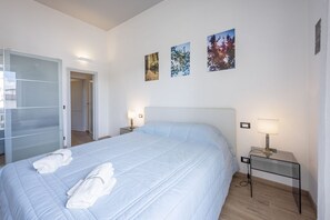 Un dormitorio con una cama, dos lámparas de noche, una mesita de vidrio y tres cuadros en la pared.