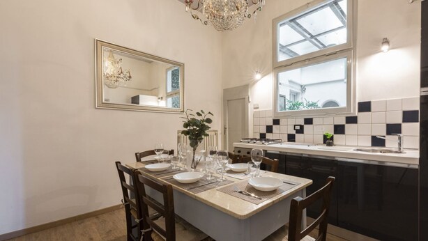 A dining area with a table set for four, a chandelier, a large mirror, and a kitchen with black and white tiles.