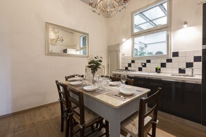Comedor con mesa para cuatro, una araña, un espejo grande y una cocina con baldosas blanco y negro.
