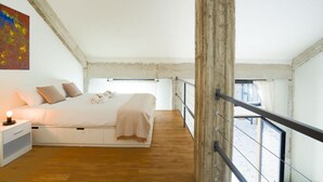 A bedroom with a large bed, wooden flooring, a nightstand, and a view through a window.