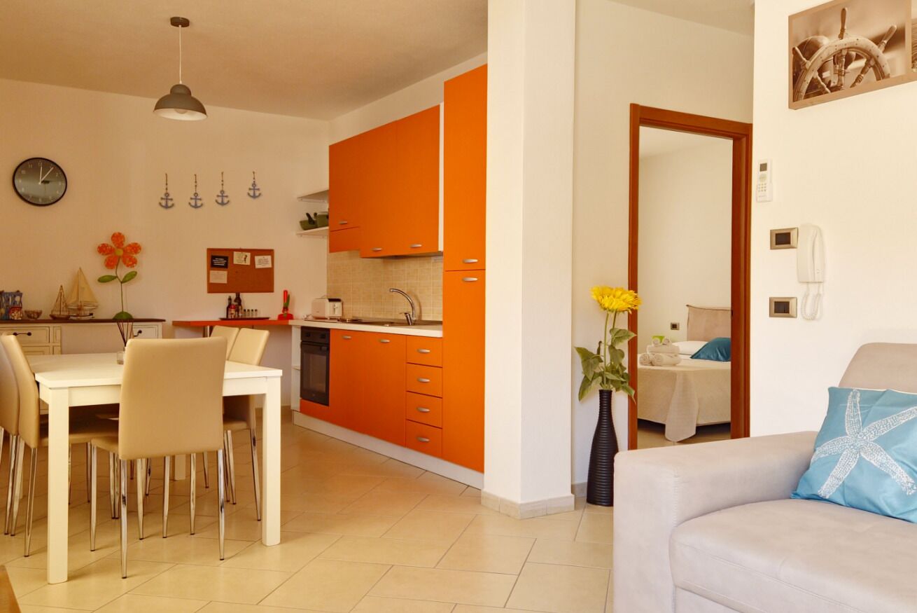 Eine moderne Küche mit orangenen Schränken, ein Essbereich mit einem weißen Tisch und Stühlen sowie ein Wohnzimmer mit einer Couch und einer dekorativen Vase.