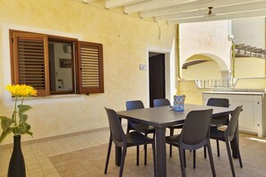 A dining area with a table and chairs, a vase with yellow flowers, and a kitchen area with a counter and a window with wooden shutters. - [Casalinda] Apartment in Orosei with veranda (Orosei)