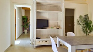 A room with a TV on a white cabinet, a dining table with chairs, and a potted plant. - [Arcada 5] Deluxe apartment with 2 bathrooms (Orosei)