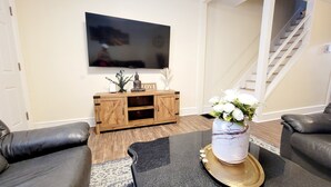 Una sala de estar con un aparador de madera, un televisor de pantalla plana montado en la pared, un florero con flores sobre una mesa y dos sofás de cuero.