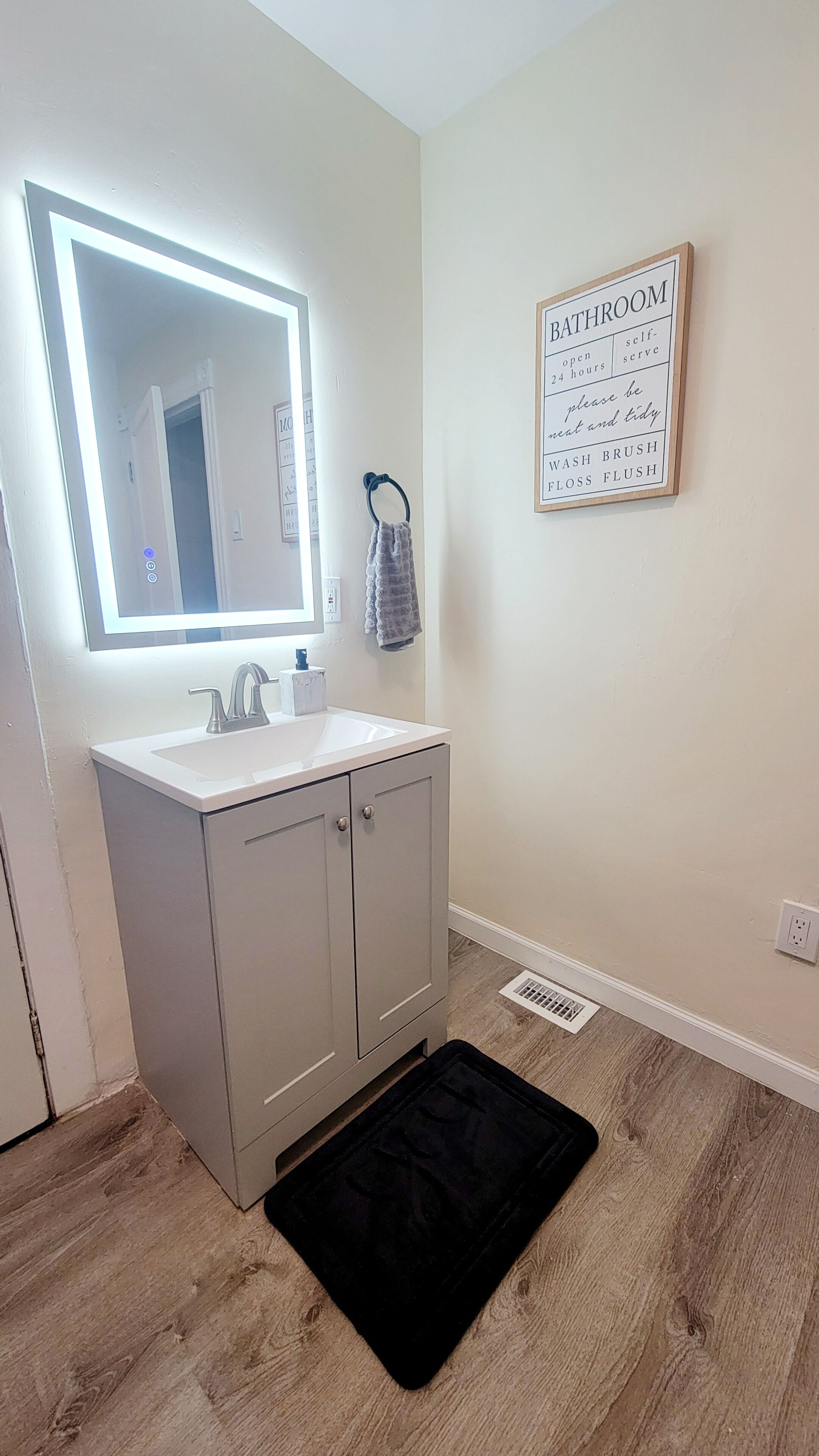 Une salle de bain avec un lavabo blanc, un miroir doté d’un éclairage intégré, un porte-serviettes et un panneau indiquant que la salle de bain est ouverte 24 heures sur 24.