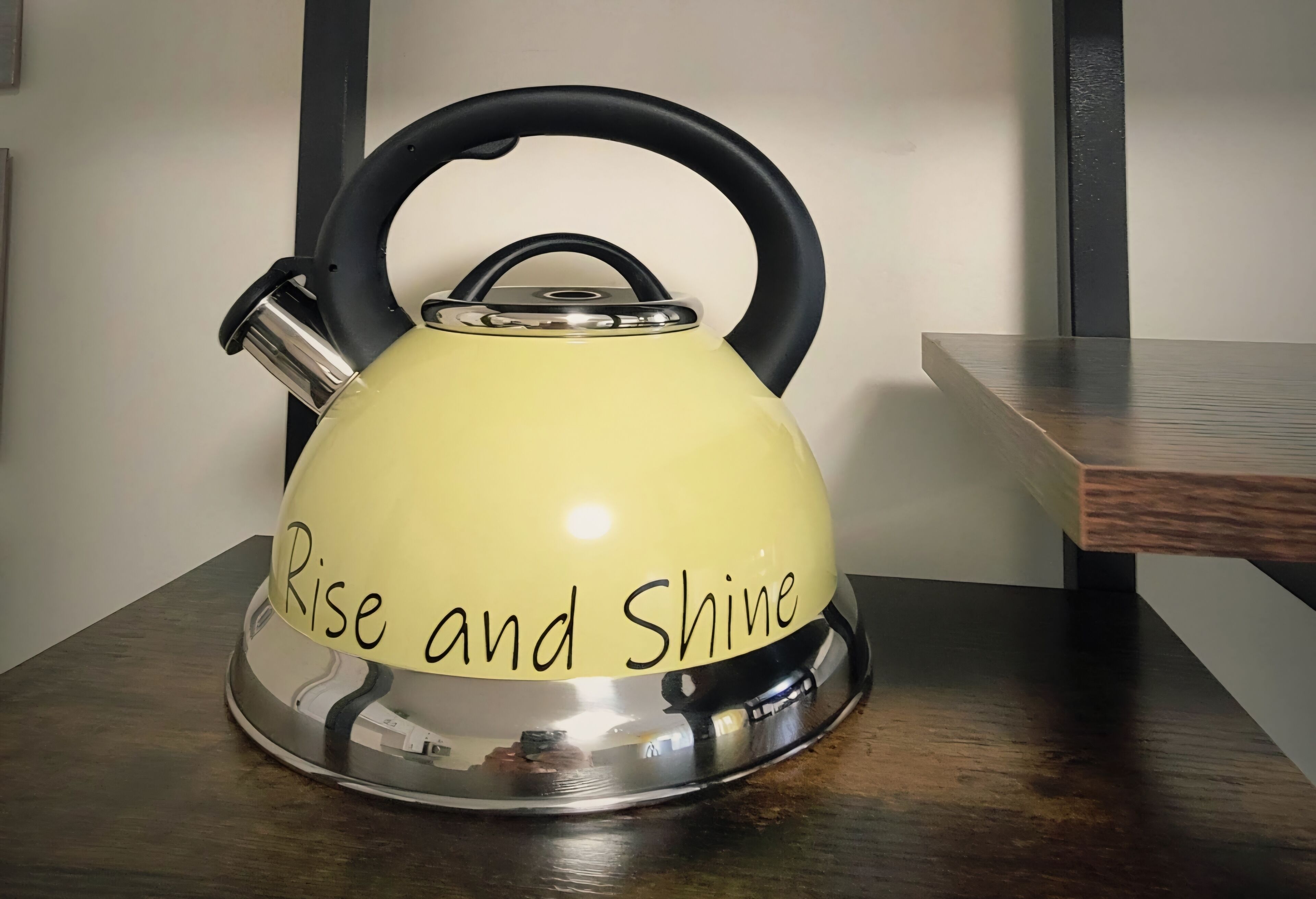 A yellow kettle with the inscription "Rise and Shine" on its base, placed on a wooden table against a wall.
