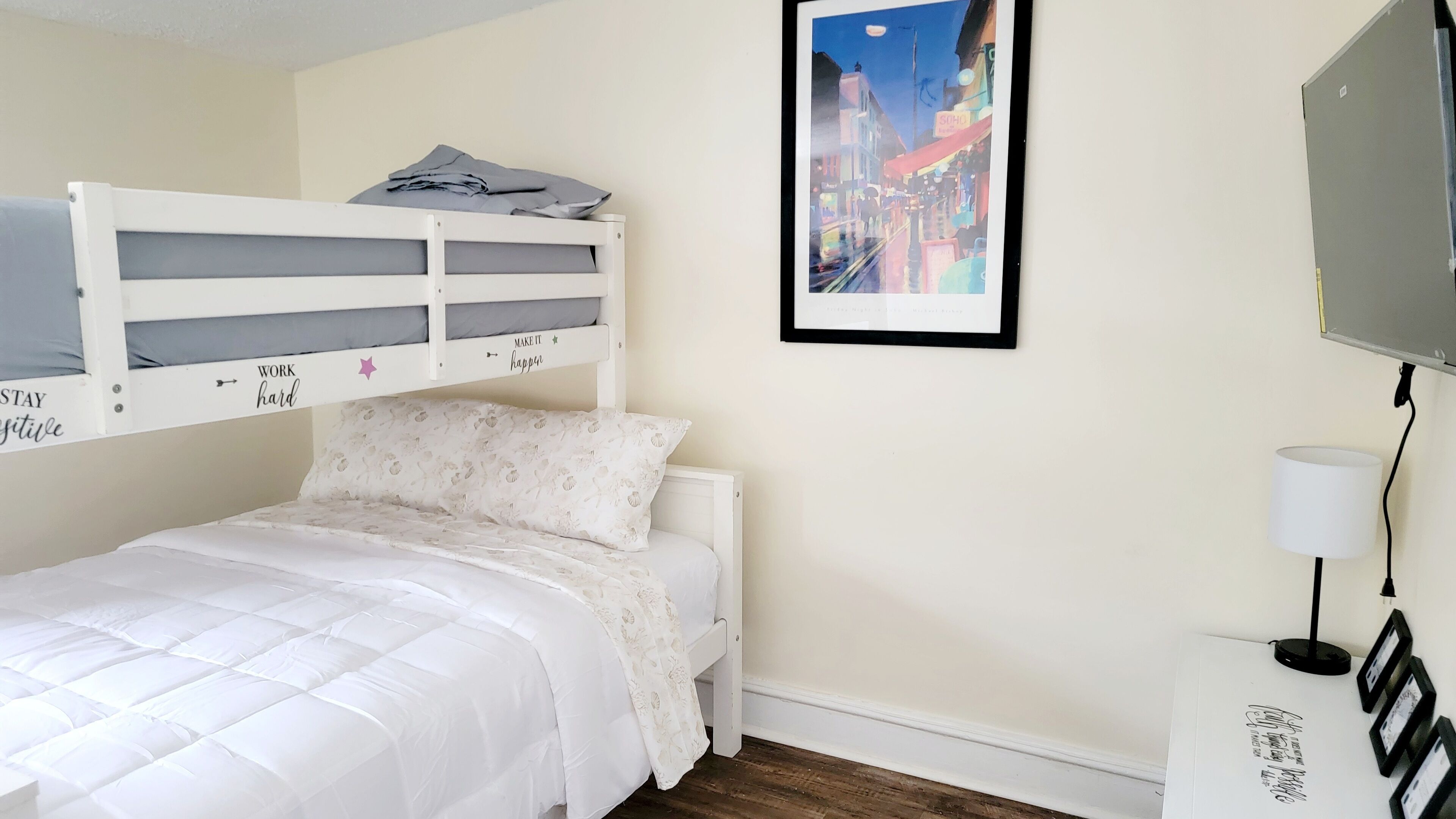 A bedroom with a bunk bed, a single bed, a framed cityscape picture, a desk with a lamp, and a framed certificate.