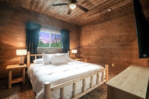 A wooden cabin bedroom with a bed, nightstands, a ceiling fan, and a window with curtains.