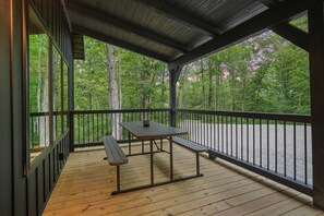 A covered outdoor deck with a picnic table and benches, surrounded by a wooden railing and overlooking a forested area. - Game Haven | Hocking Hills, Game Room, Hot Tub (McArthur)