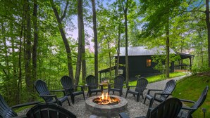 Ein rundes Feuerkörbchen, umgeben von schwarzen Adirondack-Stühlen in einer Waldlandschaft, mit einer Holzhütte im Hintergrund.