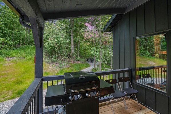 A covered outdoor area with a grill, chairs, and a view of a forested path.