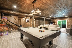 A pool table with balls, a sofa, and a hanging light fixture in a room with wooden walls and a ceiling. - Game Haven | Hocking Hills, Game Room, Hot Tub (McArthur)