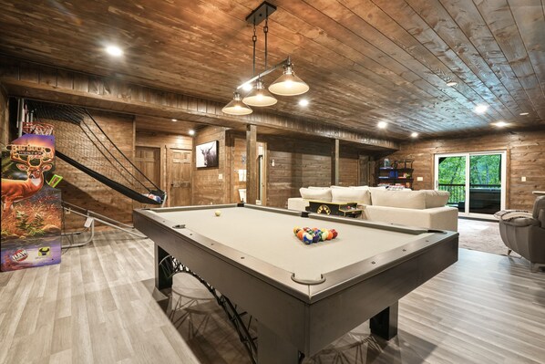 A pool table with balls, a sofa, and a hanging light fixture in a room with wooden walls and a ceiling.