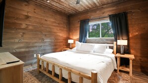 Ein Schlafzimmer einer Holzhütte mit einem Bett, Nachttischen, einem Deckenventilator und einem Fenster mit Vorhängen.