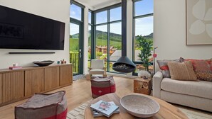 A modern living room with a large TV, a fireplace, and a view of a mountainous landscape through large windows. - Stowe Mt Resort | Spruce Peak | 4 Bed | Treehouse Condo | Sleeps 16 (Stowe)