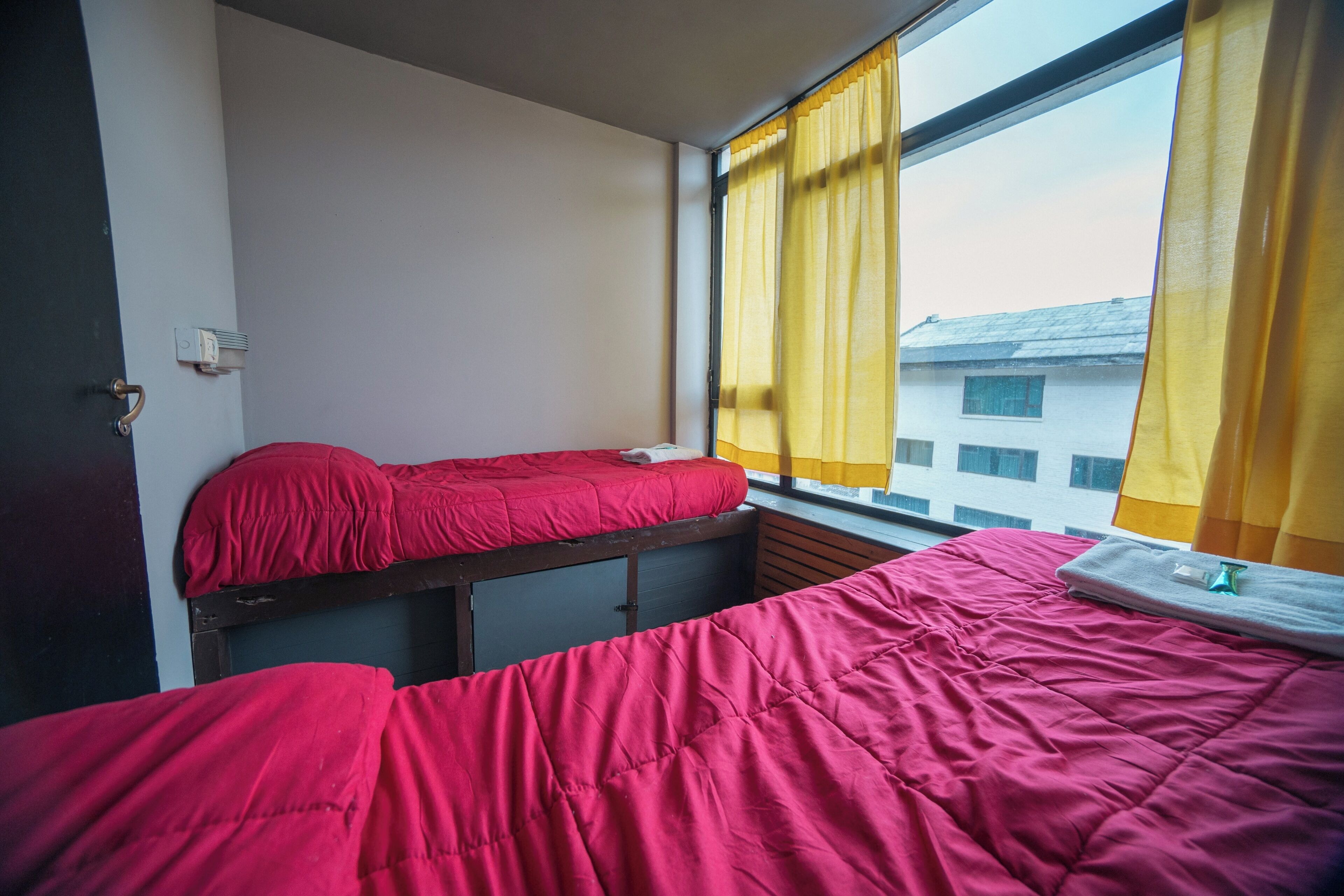 A room with two beds, each with a red bedspread, a window with yellow curtains, and a view of buildings outside.