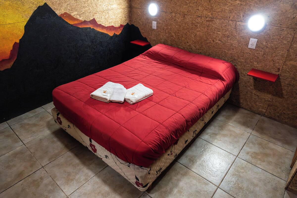 Una cama con una colcha roja, un colchón estampado con motivos florales y dos toallas.