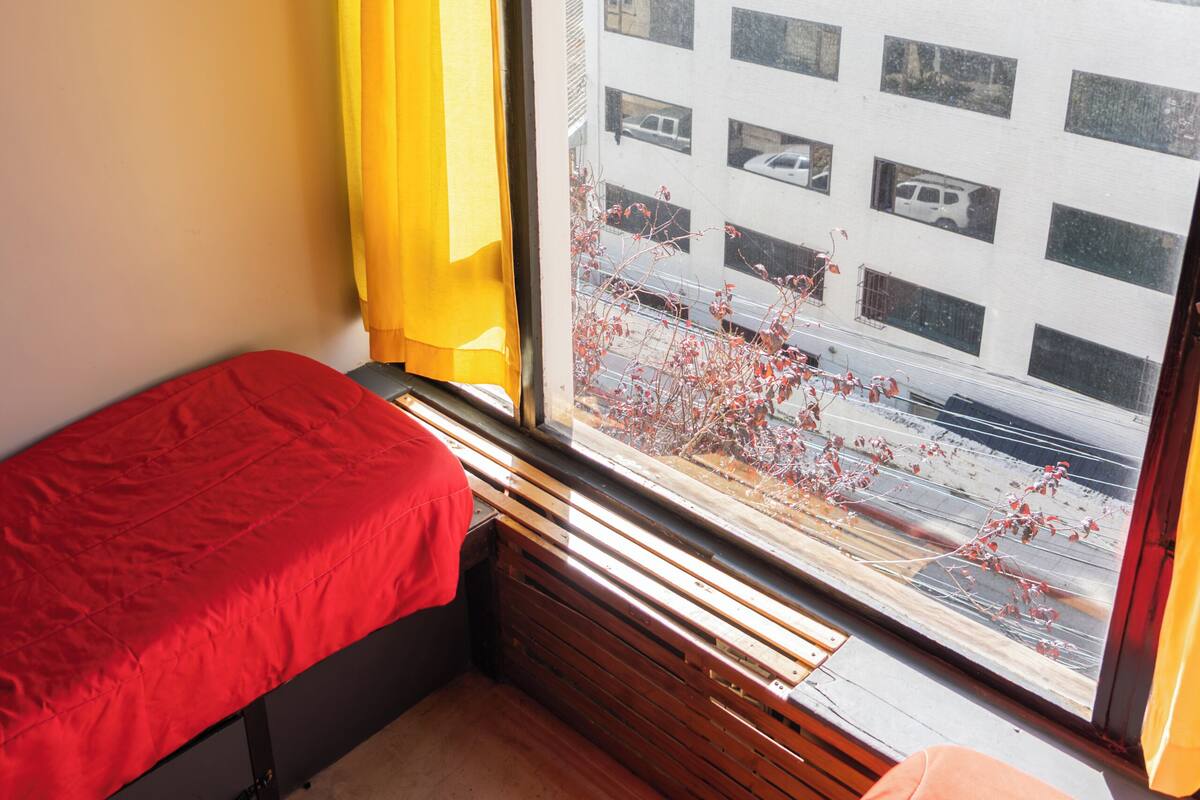 Una habitación pequeña con dos camas, una ventana con cortinas amarillas y vistas a un edificio.