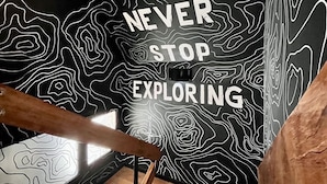 Una escala al costat d'una paret negra amb línies topogràfiques blanques i el text "NEVER STOP EXPLORING".