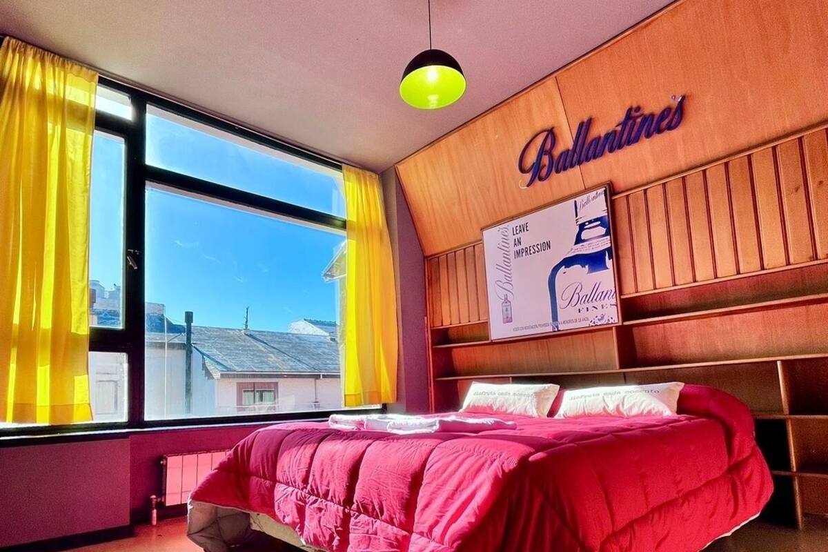 Un dormitorio con una cama, una ventana con cortinas amarillas, un cabecero de madera con un cartel, y vista a un puente.