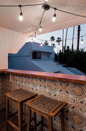 An outdoor bar area with a patterned wall, two wicker stools, and string lights.