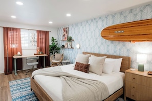 Een slaapkamer met een bed, nachtkastje, bureau, stoel en een surfplank aan de muur.