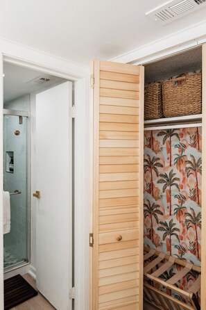 A walk-in closet with a sliding wooden door, a patterned shower curtain, and wicker baskets on shelves. - NEW Desert Escape: Patio Bar, Pool View, 3 King Beds (Palm Springs)
