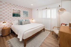 A bedroom with a bed, bedside lamps, a wooden bedside table, a chair, and a window with curtains. - NEW Desert Escape: Patio Bar, Pool View, 3 King Beds (Palm Springs)