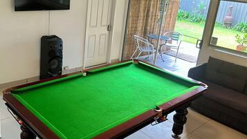 A pool table in a room with a television mounted on the wall, a door, and a view of a garden through a sliding door.
