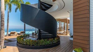 Un espacio exterior moderno con una estructura negra curva, terraza de madera y sillones de descanso junto a una piscina con vista al océano.
