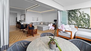 Habitación de hotel moderna con zona de comedor, sofá, mesa baja con una orquídea en maceta y vistas al exterior.